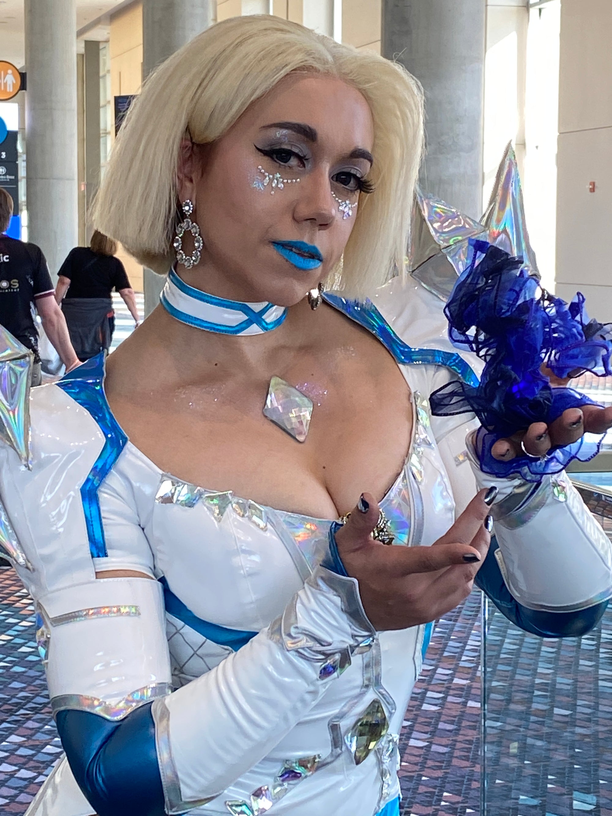 A woman in a white and blue futuristic costume with holographic accents, holding a blue tulle bouquet in an indoor public space.