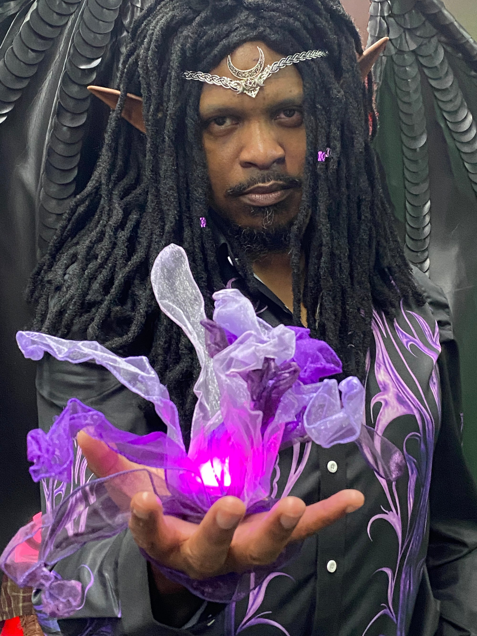A man wearing a dark fantasy elf costume with long black dreadlocks, pointed ears, and a silver crescent moon headpiece, holding a glowing purple magical orb in his hand.