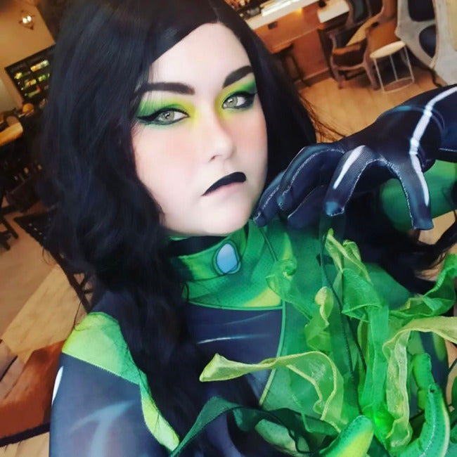 A close-up selfie of a person in a green and black costume with dramatic green eye makeup, holding a green leafy prop in an indoor setting.